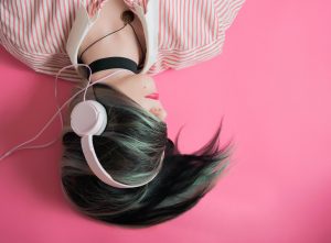 Image of woman with dark hair laying on a pink floor wearing pink headphones. Her face is covered by her hair